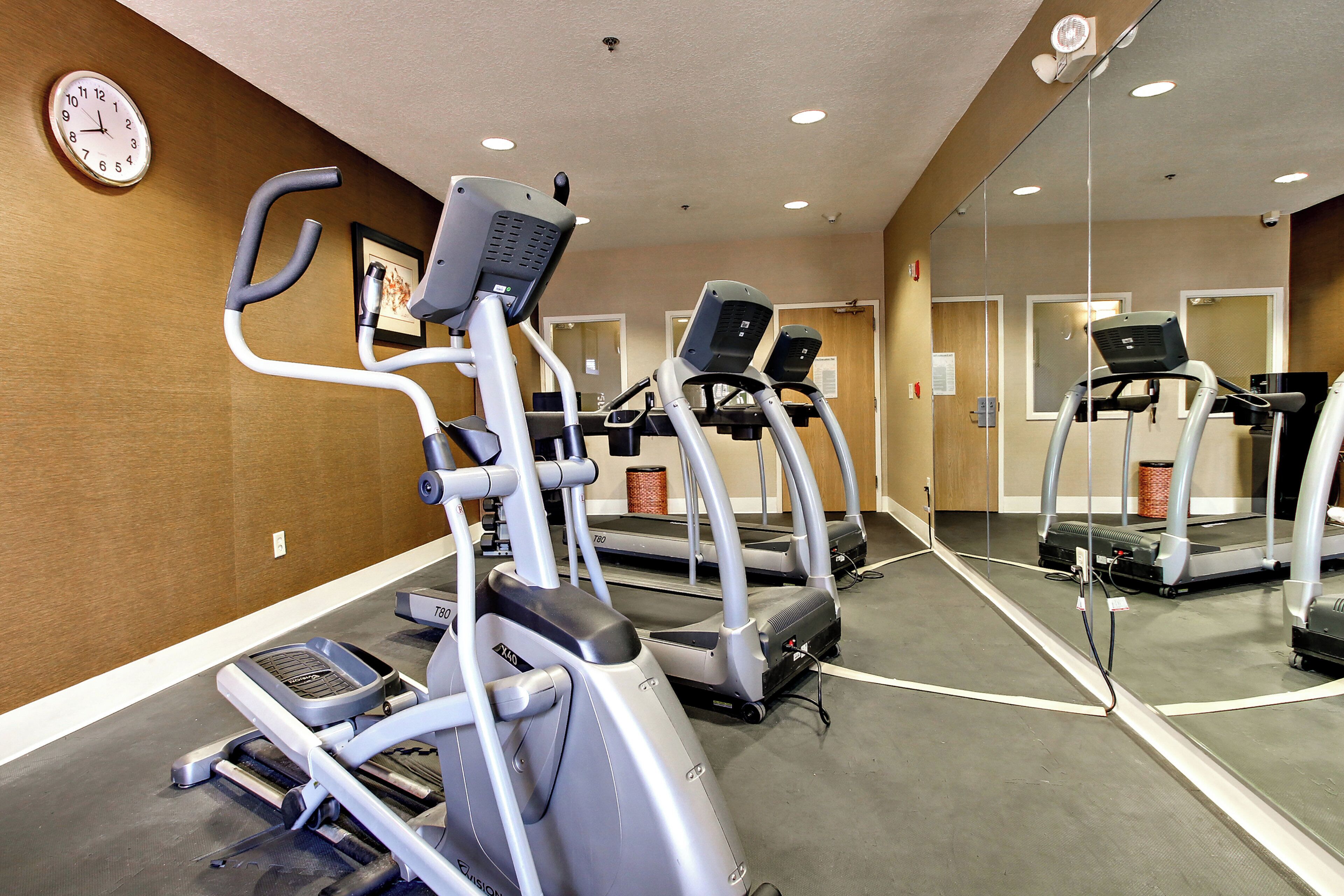 Une salle de sport équipée de tapis de course et de machines elliptiques, avec une horloge au mur et un miroir qui reflète l’équipement.