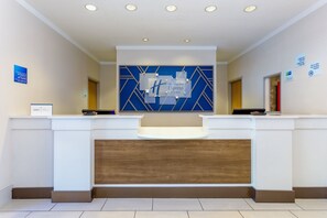 Una recepción de hotel con diseño moderno, que presenta una paleta de colores azul y blanco, un panel de acento de madera y una pantalla digital sobre el mostrador.