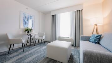 A modern hotel room with a white sofa, a white ottoman, a small table with chairs, and a large window with curtains.