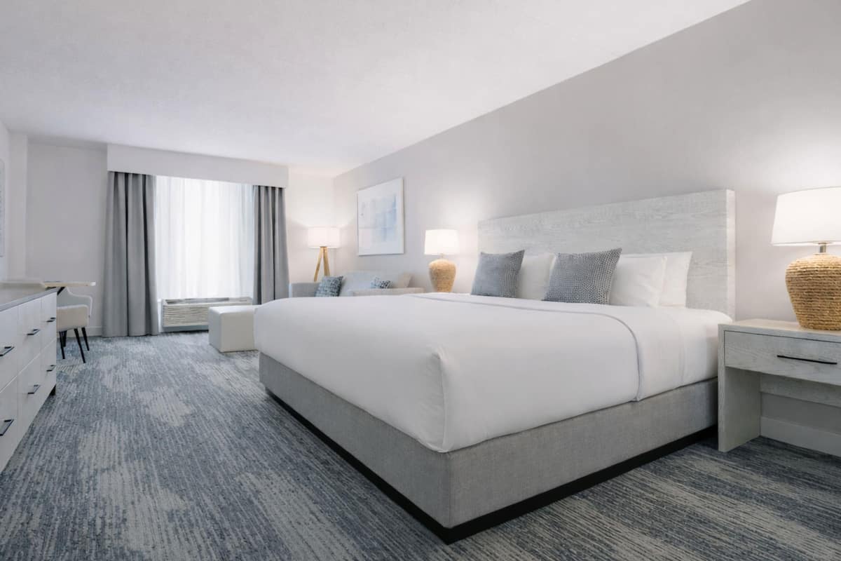 A modern hotel room with a large bed, a desk with a flat-screen TV, a seating area with a chair, and a window with curtains.