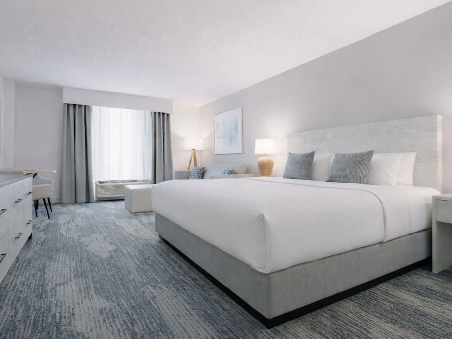 A modern hotel room with a large bed, a desk with a flat-screen TV, a seating area with a chair, and a window with curtains.