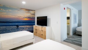 A bedroom with a large wall mural of a sunset over the ocean, a TV on a wooden cabinet, and a bed with a white mattress. - Hotel Bethany Beach (Bethany Beach)