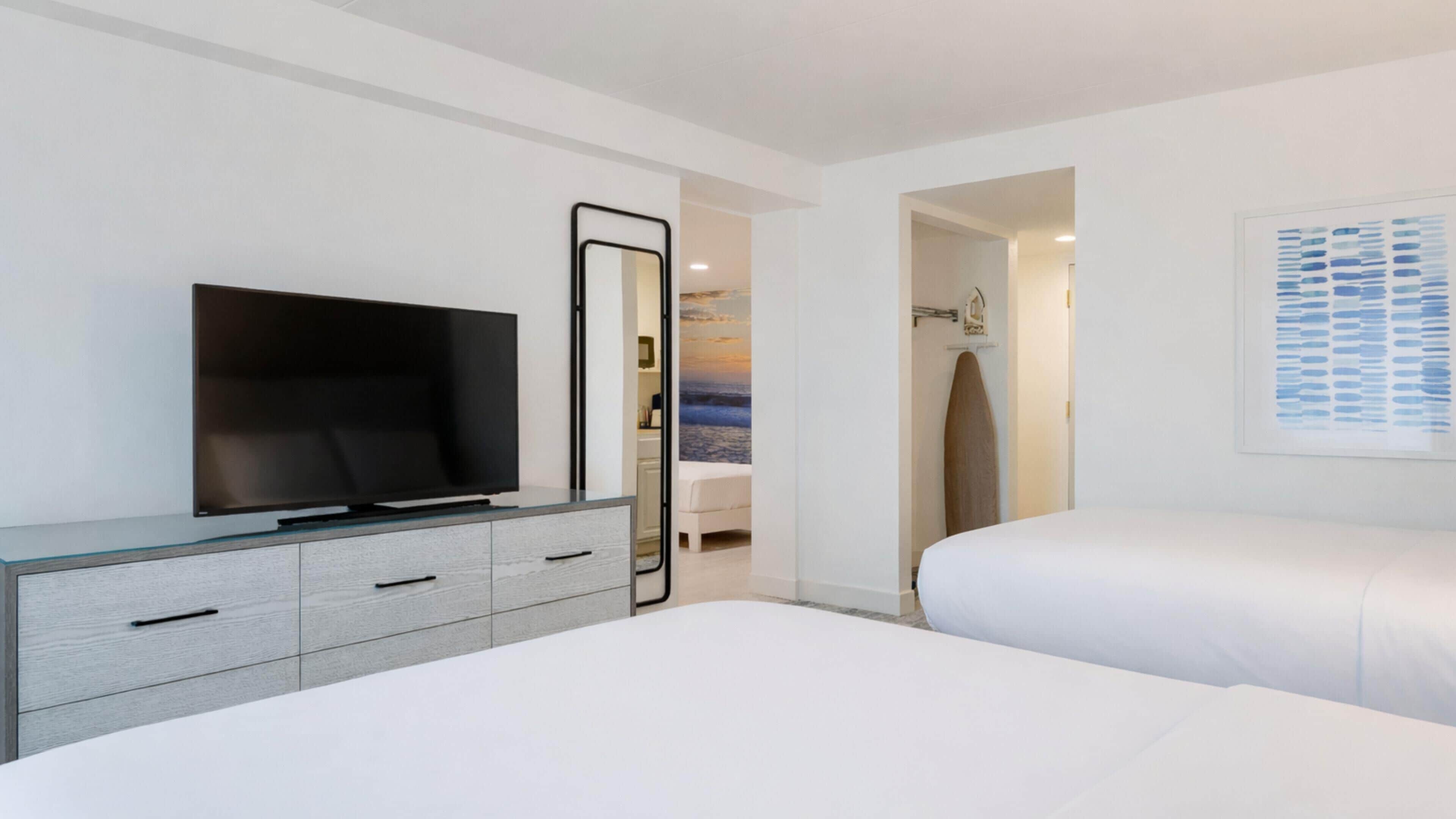 Une chambre d’hôtel moderne avec un téléviseur à écran plat posé sur une console grise, un grand lit avec une literie blanche, une œuvre d’art encadrée au mur et une porte donnant accès à la salle de bain.