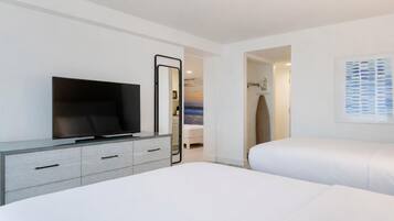 Une chambre d’hôtel moderne avec un téléviseur à écran plat posé sur une console grise, un grand lit avec une literie blanche, une œuvre d’art encadrée au mur et une porte donnant accès à la salle de bain.