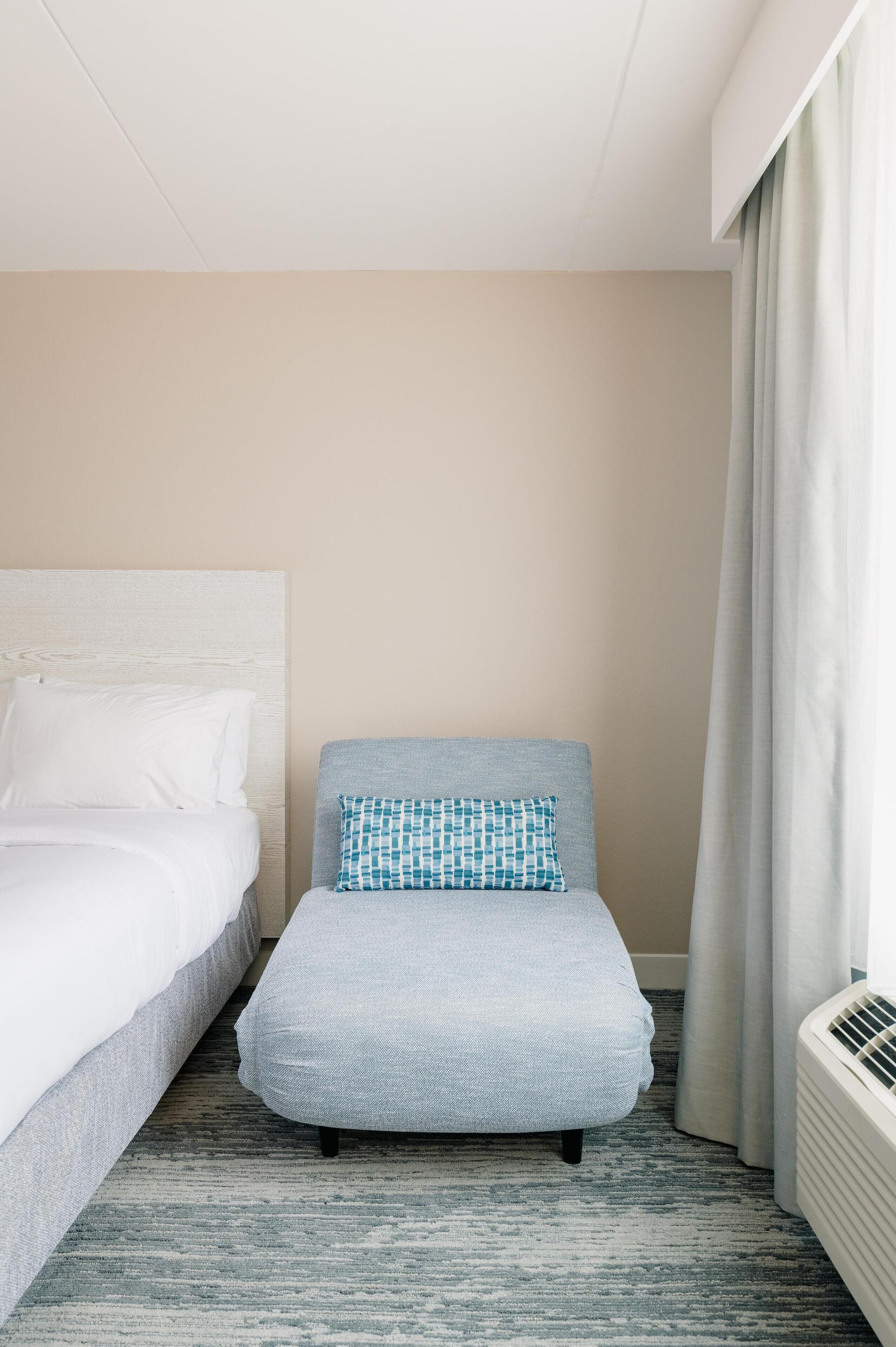 Une chambre d’hôtel avec un lit, un petit coin salon avec un coussin, un sol recouvert de moquette et une fenêtre avec des rideaux.