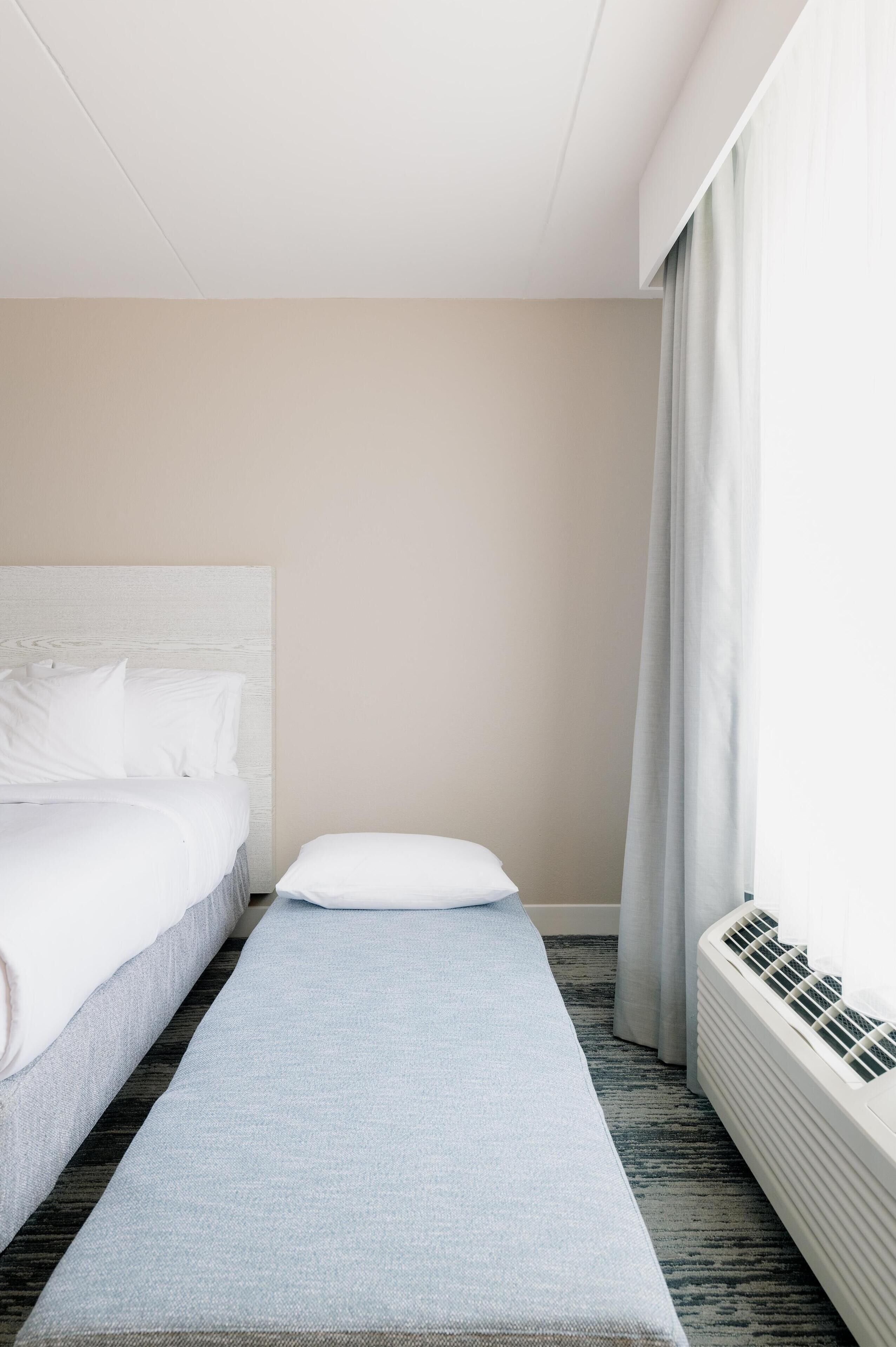 A hotel room with a bed, a bench, a window with curtains, and a wall-mounted air conditioner.