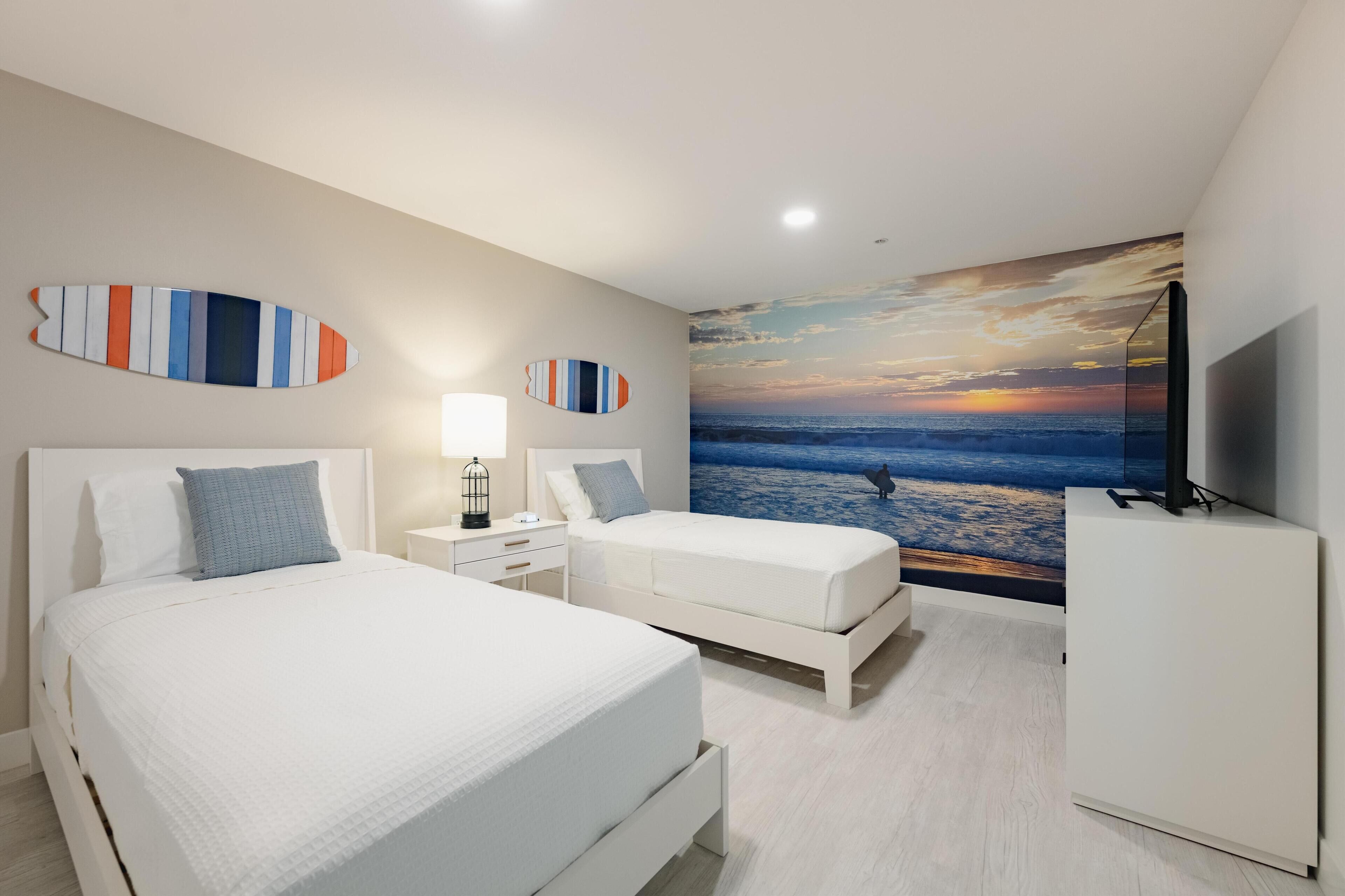 A modern hotel room with two beds, a large wall mural of a sunset over the ocean, and a flat-screen TV on a cabinet.