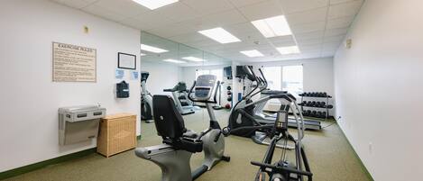Une salle de sport équipée de machines d’exercice, dotée d’un mur couvert de miroirs et d’un panneau d’affichage où sont affichées les règles.