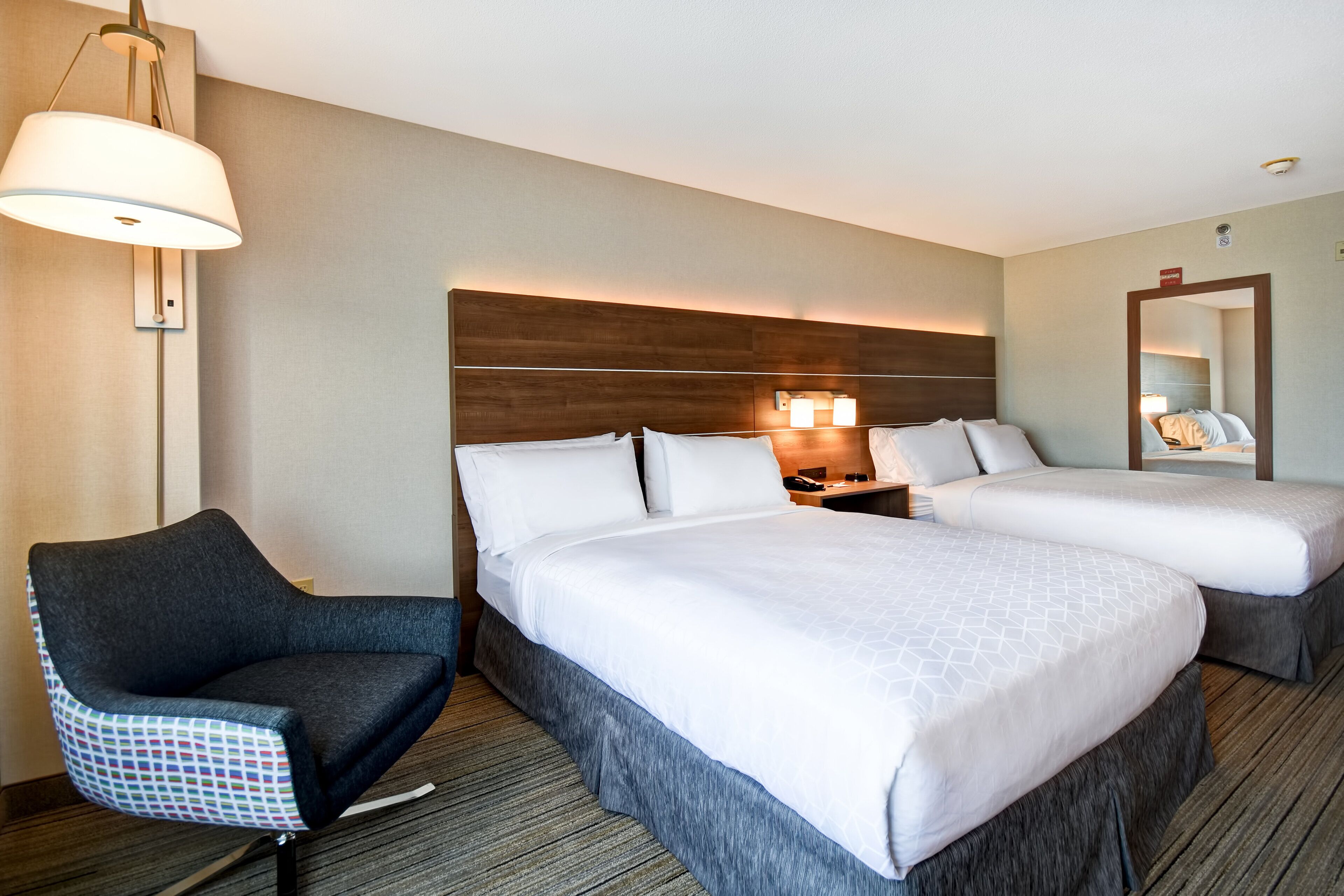 Habitación de hotel con dos camas, una silla, una mesita de noche con lámpara y un espejo.
