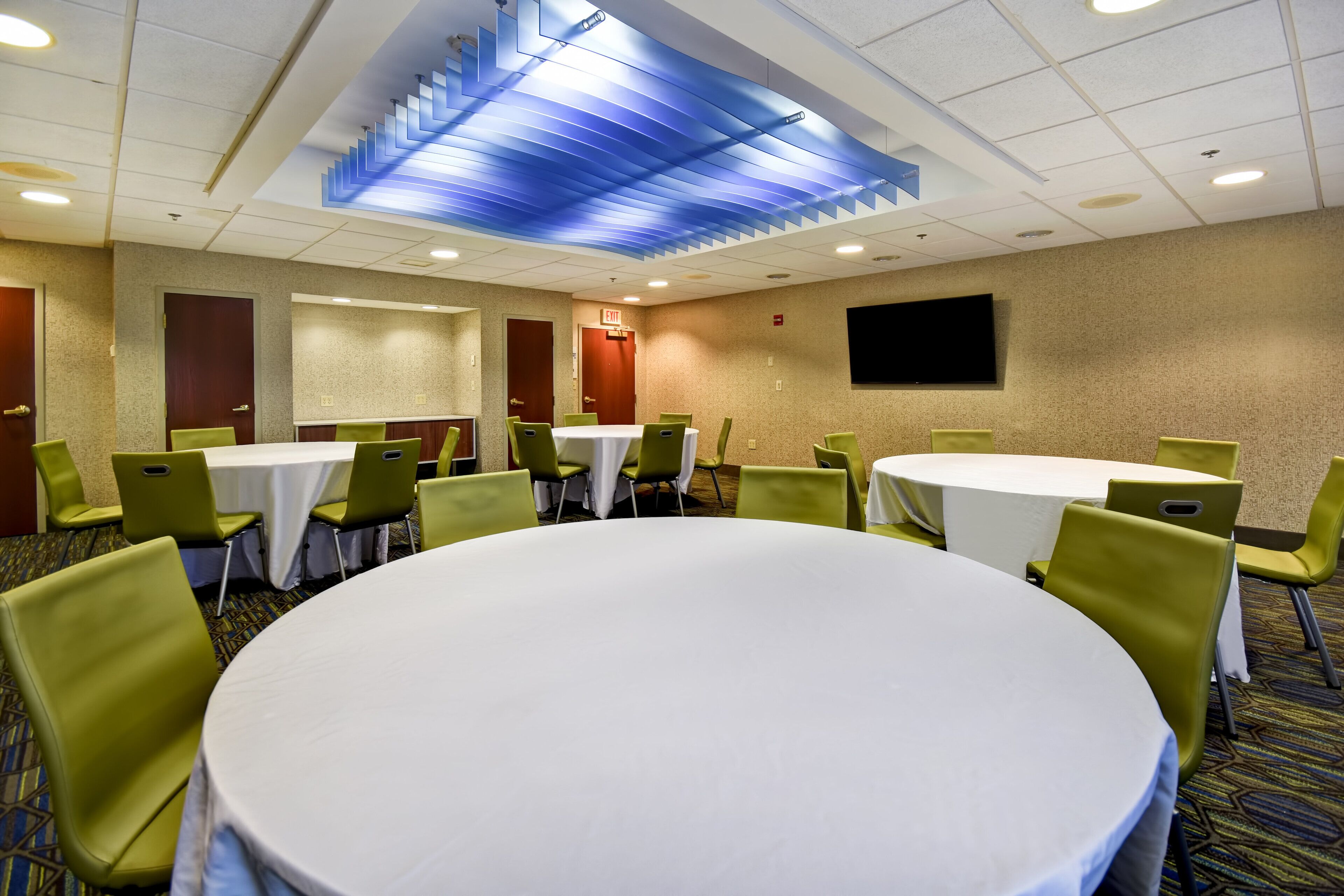 A conference room with round tables and green chairs, a flat-screen TV on the wall, and a blue ceiling design.