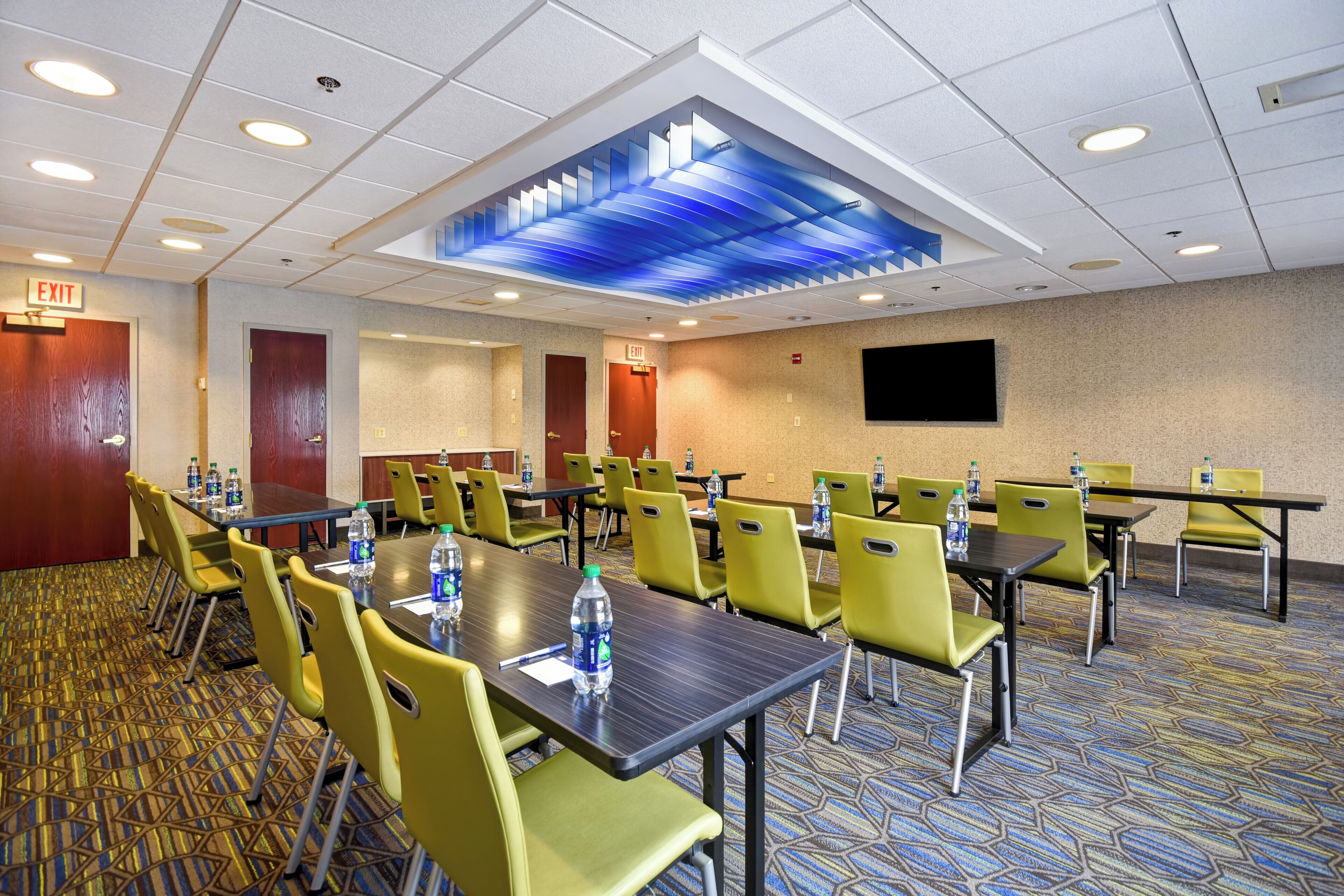 A conference room with multiple tables and chairs, equipped with water bottles, and an exit sign on the wall.