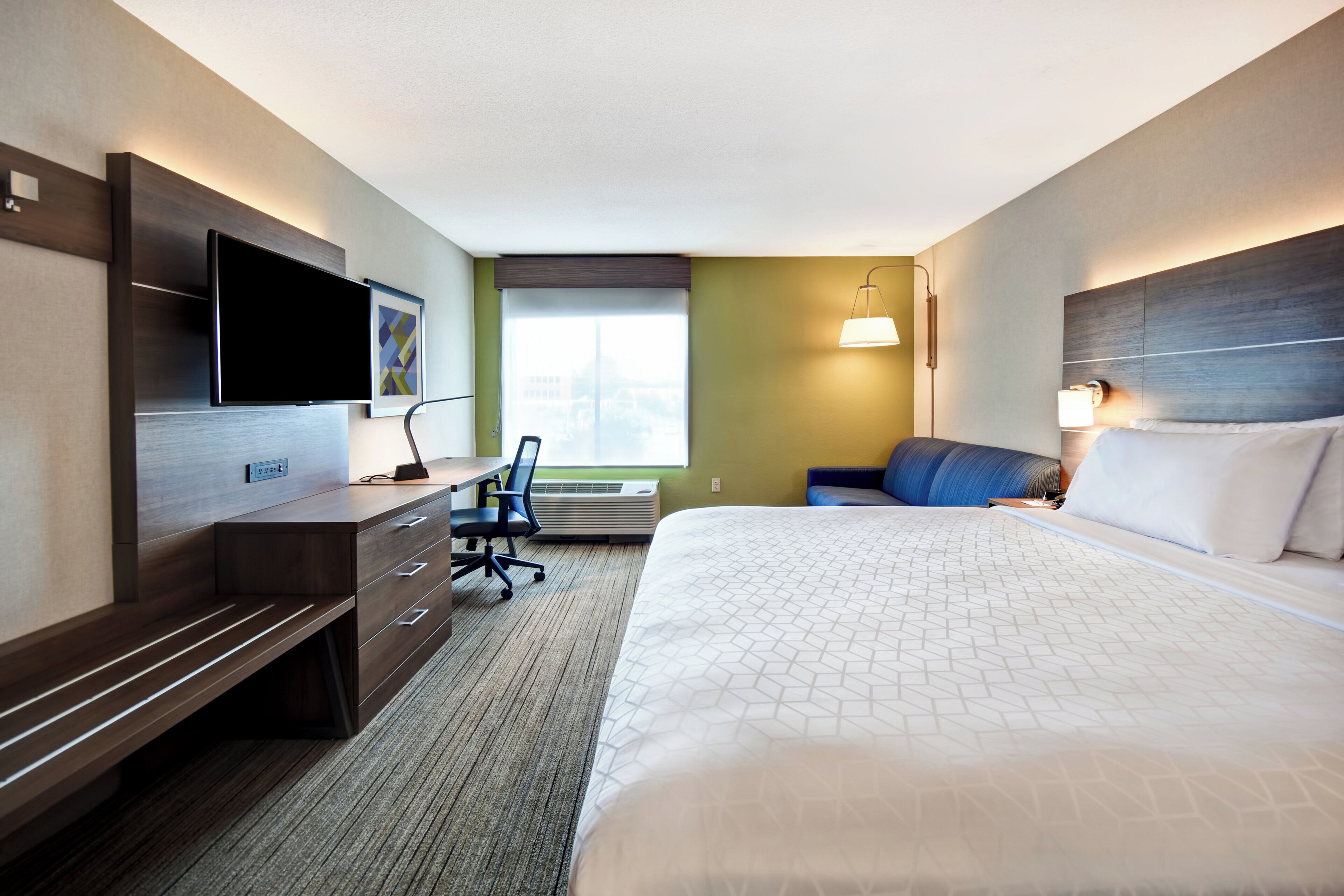 Una habitación de hotel con cama, escritorio, silla, televisor y ventana.