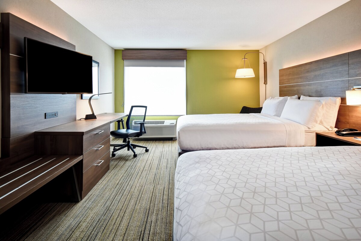A hotel room with a flat-screen TV, a desk with a chair, two beds, and a large window.