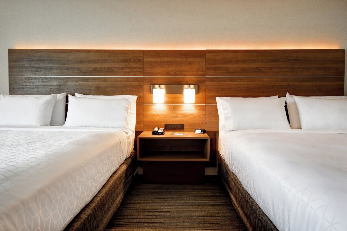 A hotel room with two beds, a wooden headboard, bedside tables with lamps, and a small nightstand.