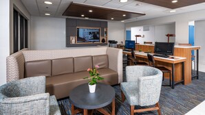 Un hall d’hôtel moderne avec un canapé en cuir marron, deux fauteuils à motifs, une table ronde agrémentée d’une plante en pot, un bureau avec un ordinateur et un téléviseur fixé au mur.