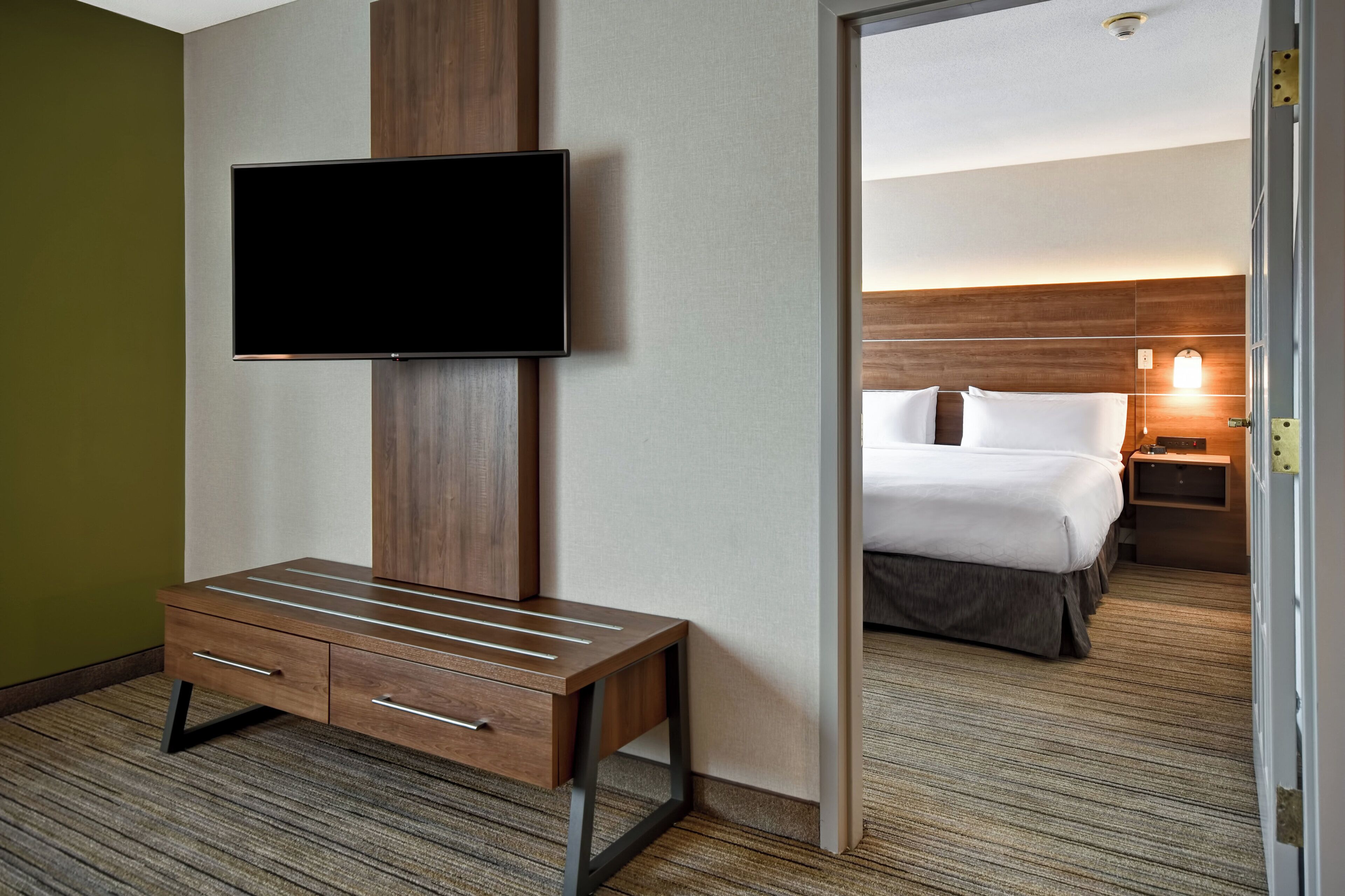 Habitación de hotel con televisor de pantalla plana en la pared, un escritorio de madera con cajones, cama con ropa blanca y mesita de noche con lámpara.