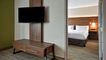 Quarto de hotel com televisão de tela plana na parede, escrivaninha de madeira com gavetas, cama com roupa de cama branca e mesa de cabeceira com abajur.