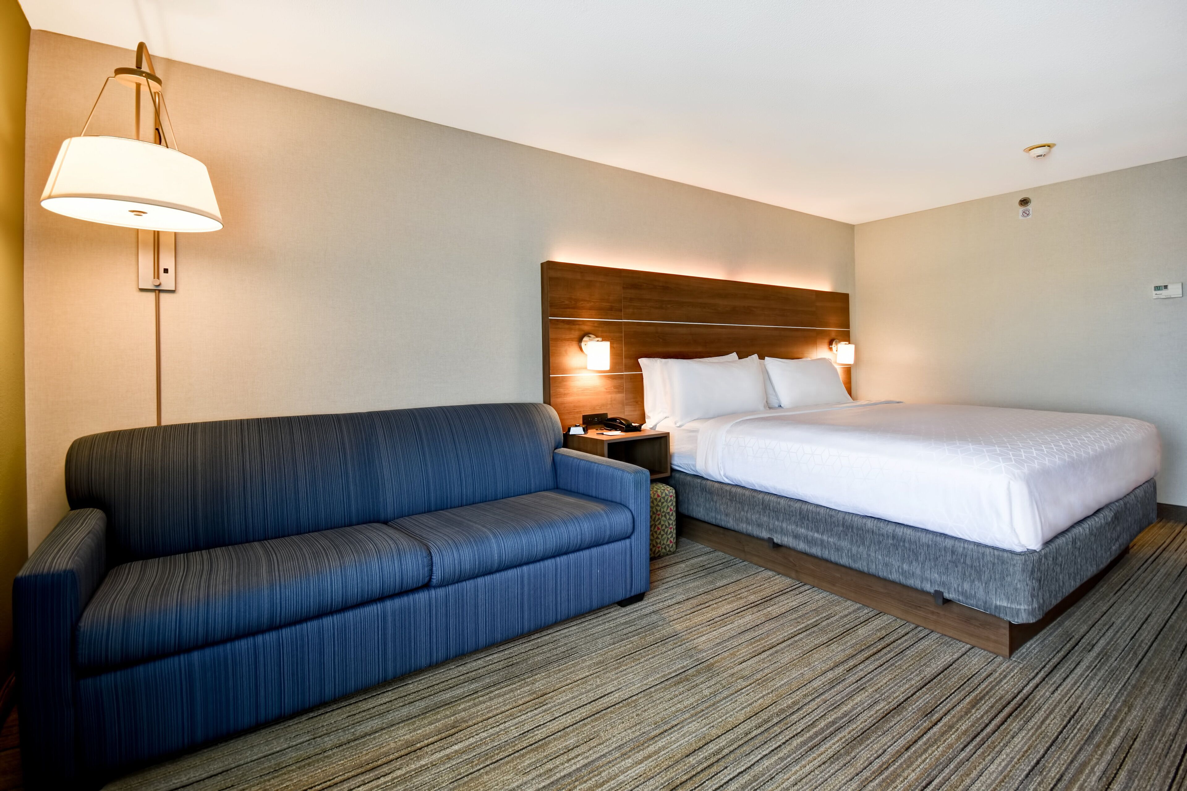 A hotel room with a bed, a sofa, a nightstand, and a wall-mounted lamp.
