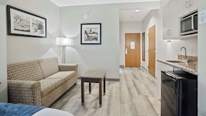 A compact hotel room with a sofa, a small table, a lamp, and a kitchenette with a microwave. - Blue Water Inn & Suites, BW Signature Collection (Sneads Ferry)