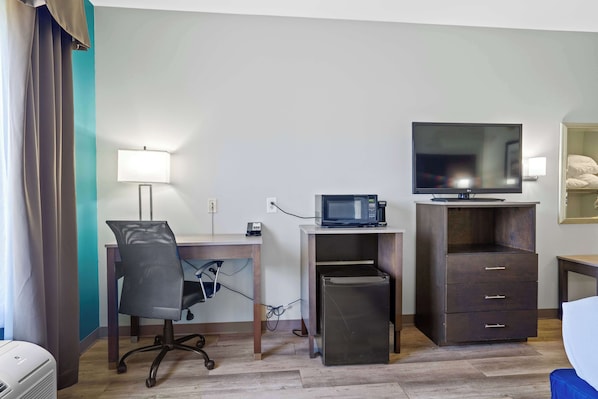 A hotel room with a desk, chair, microwave, TV, and a small refrigerator.