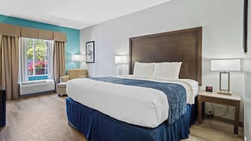 A hotel room with a large bed, a window with curtains, a chair, a bedside table with a lamp, and a wall-mounted TV.