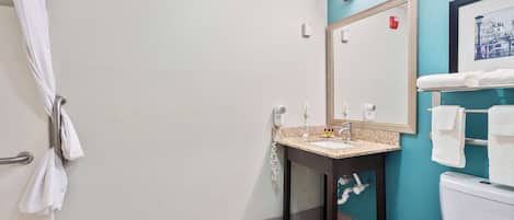 Et badeværelse med vask, spejl og toilet. Der er håndklæder hængende på væggen og en badeforhæng.