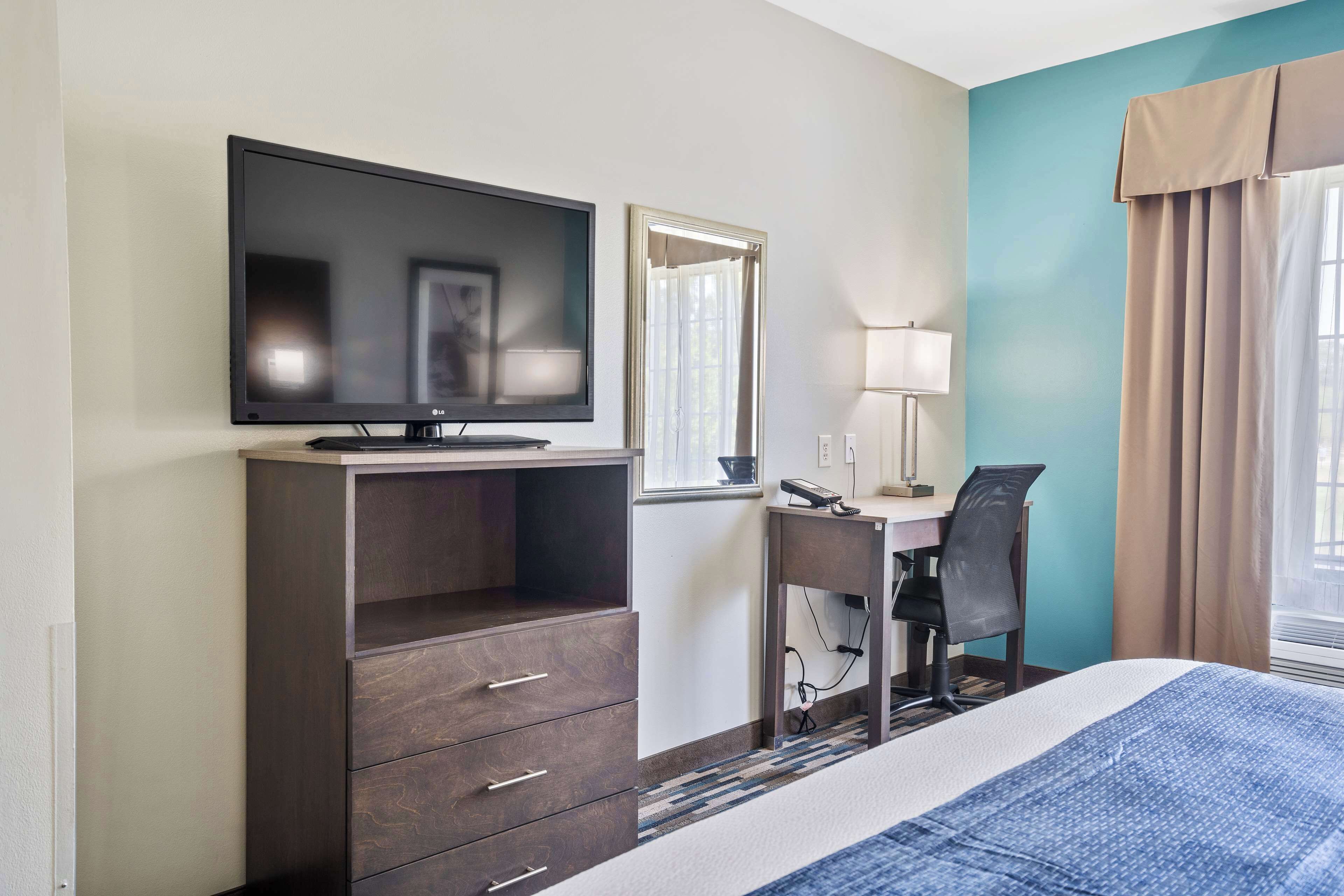 A hotel room with a bed, a desk with a chair, a television, a mirror, and a window with curtains.