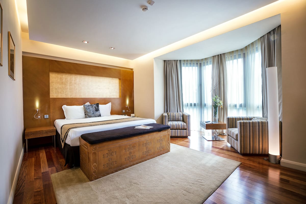 A modern hotel room with a large bed, a wooden bench, a striped armchair, and a large window with curtains.