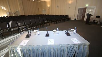 A conference room with rows of chairs, a table with microphones and water bottles, and a podium.