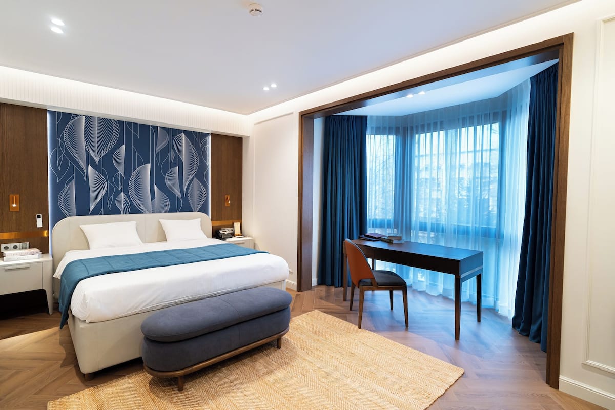 A modern hotel room with a large bed, a desk, and a view of the city through sheer curtains.