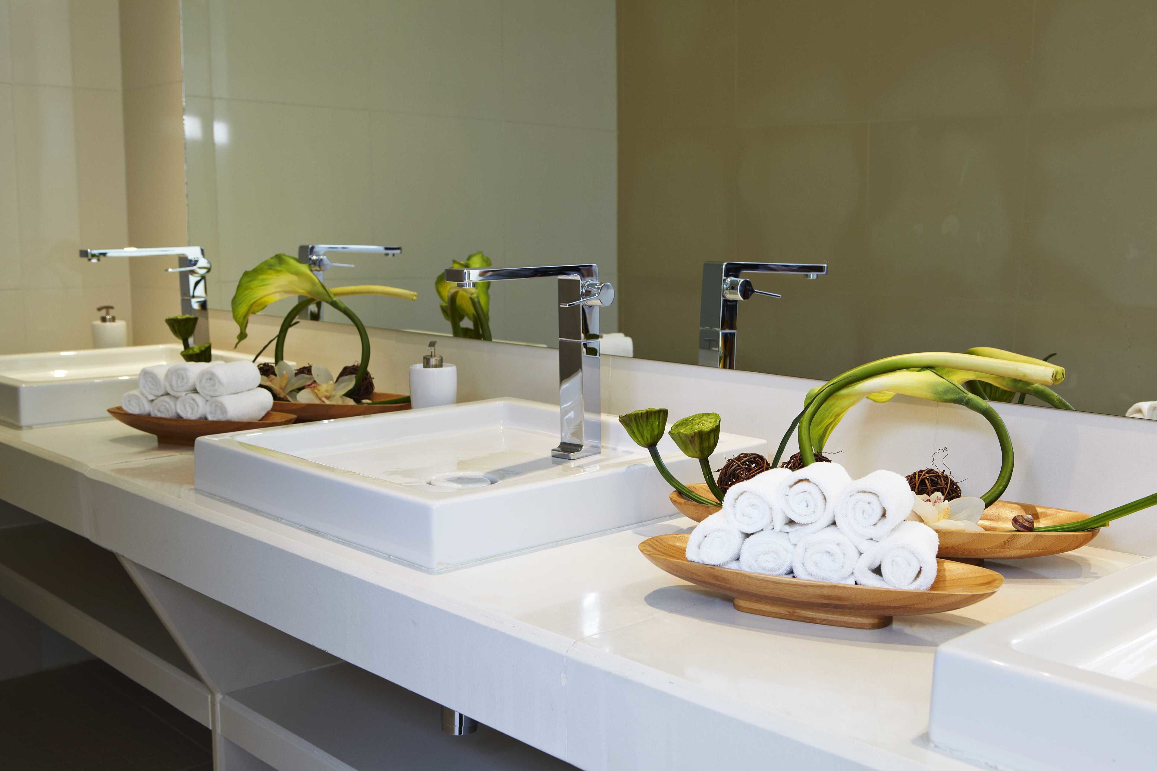 A bathroom with two sinks, each with a mirror above. There are wooden bowls with rolled towels and green leaves on the counters.