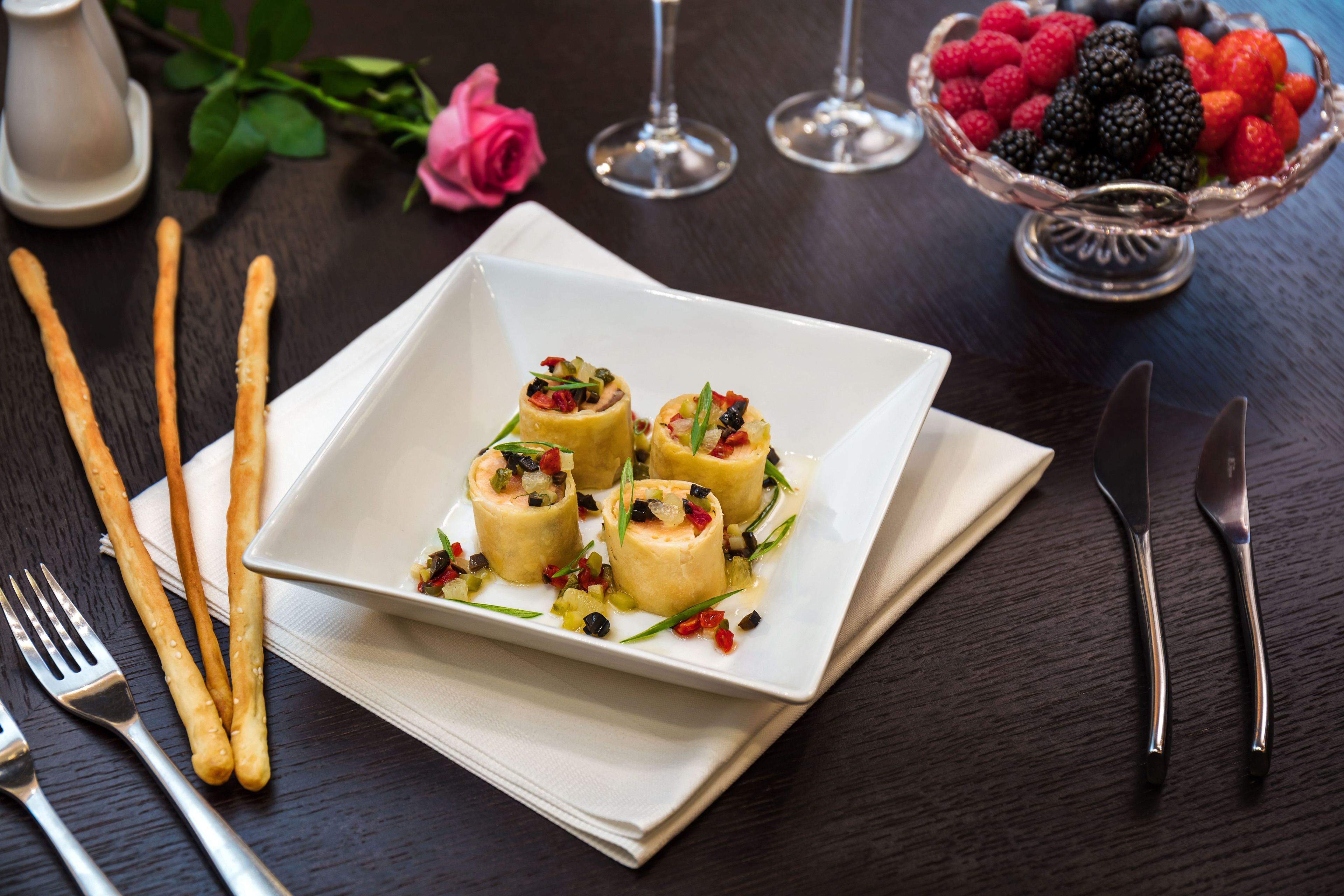 A square white plate with four rolled appetizers garnished with herbs, accompanied by a bowl of mixed berries and a glass of wine on a dark wooden table.