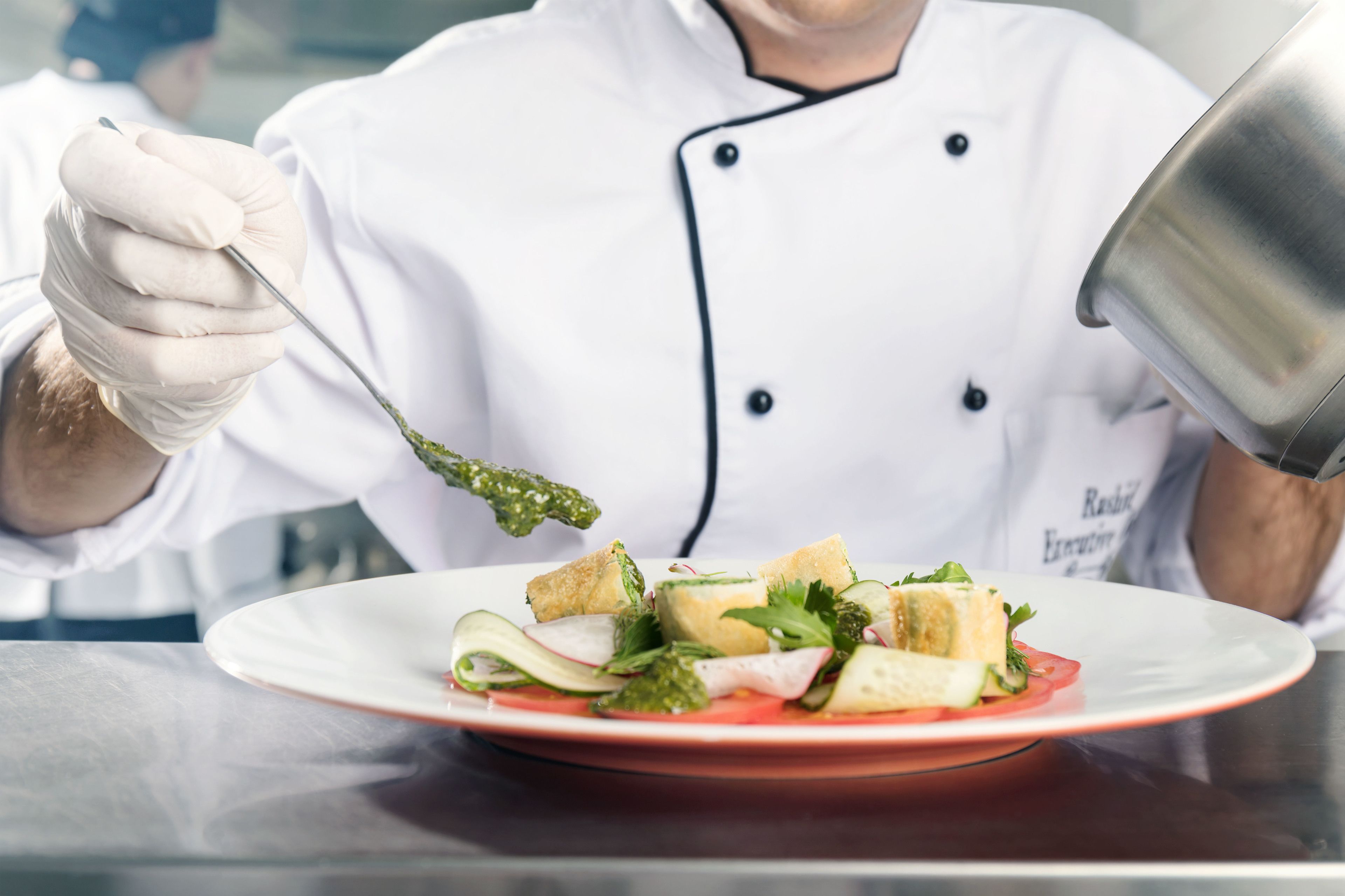 Un chef, vêtu d’un uniforme blanc, tient une cuillère au-dessus d’une assiette contenant une salade.