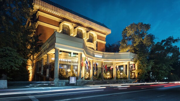 A grand hotel with a classical facade, multiple flags, and a well-lit entrance. - The Dostyk Hotel (Almaty)
