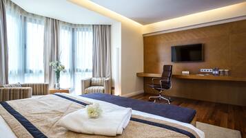 Habitación de hotel con una cama grande, un escritorio con silla, televisor montado en la pared y una ventana con cortinas.