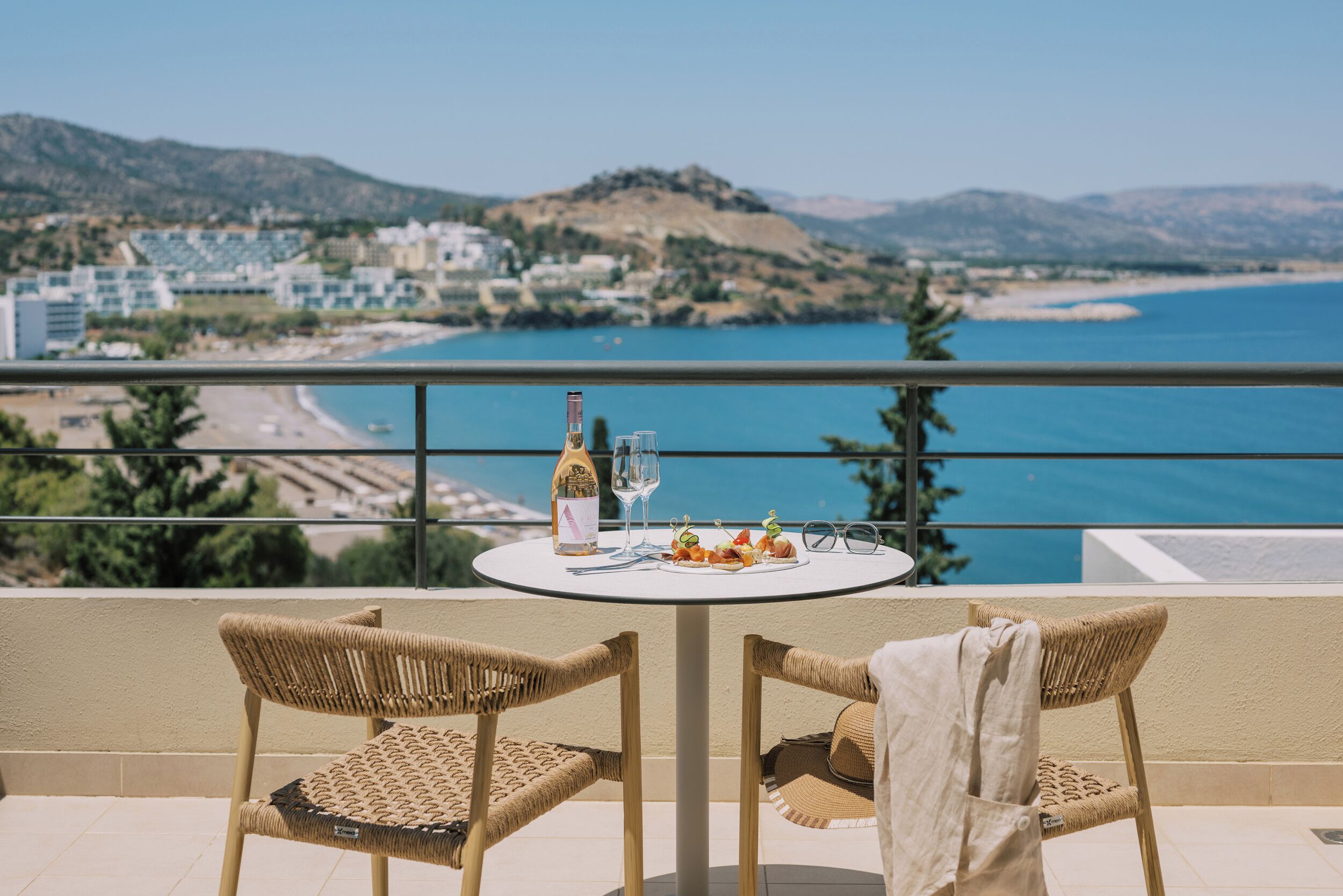 Ein Balkon mit Korbstühlen, einem Tisch mit Flasche, Gläsern und einem Teller Essen, mit Blick auf eine Küstenlandschaft mit Gebäuden und Hügeln.
