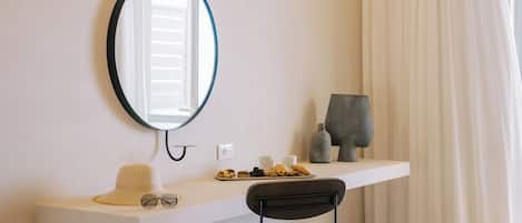 A room with a white vanity, a round mirror, a hat, glasses, a tray with pastries, and a sculpture on a shelf.