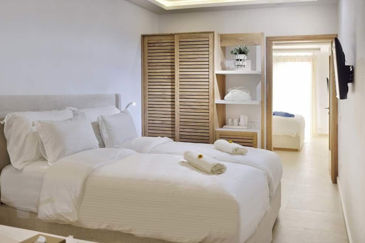 Ein Hotelzimmer mit zwei Betten, einem Holzschrank, einer kleinen Sitzecke und Blick auf das Nachbarzimmer.