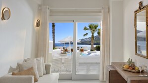 A modern hotel room with a sofa, a dining table with fruit and glasses, and a view of the beach and ocean through the glass door. - Nissaki Boutique Hotel (Mykonos)