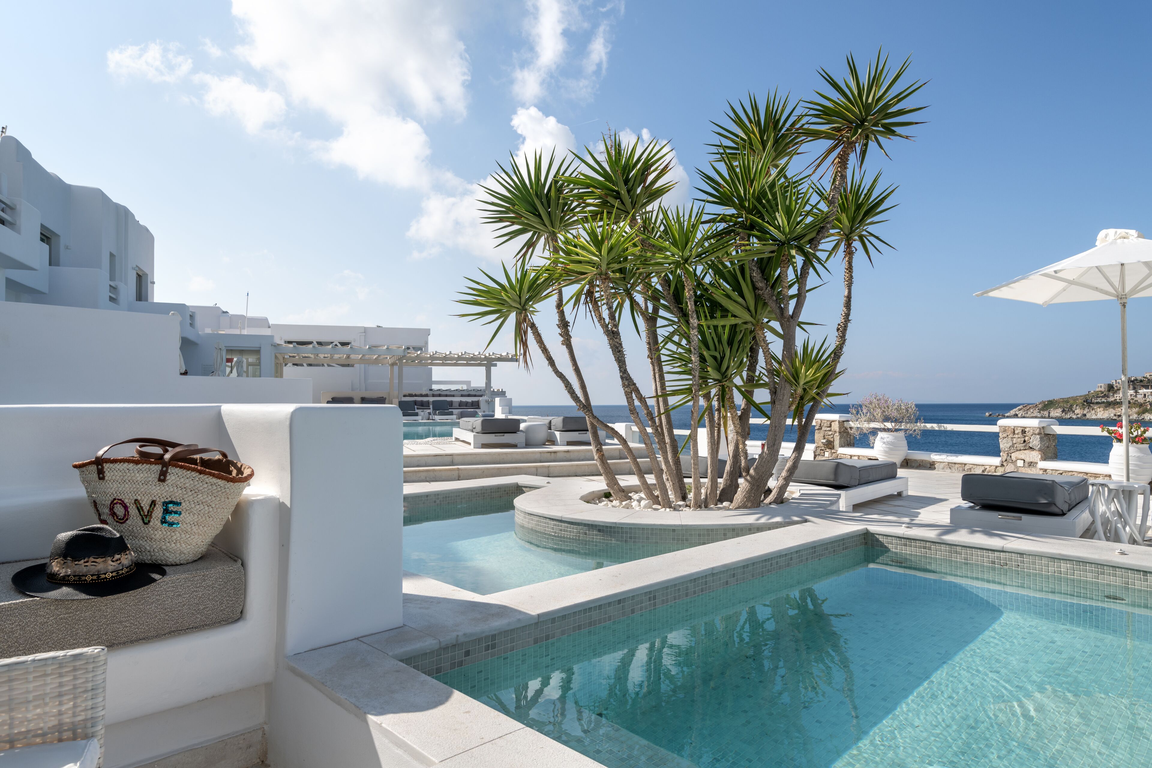 A pool area with a view of the sea, surrounded by palm trees and white buildings.