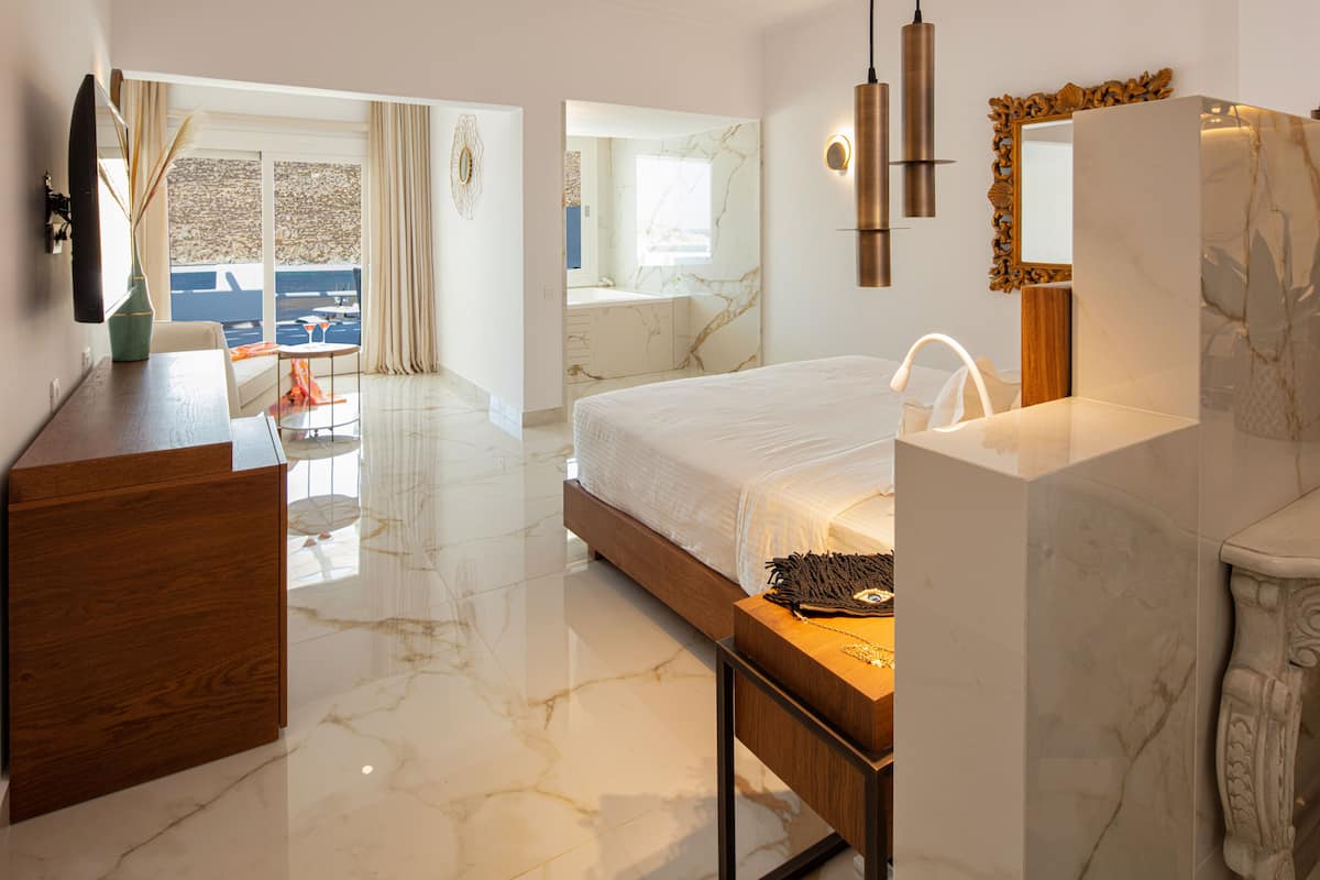 Ein modernes Hotelzimmer mit Marmorboden, einem großen Bett, einer Holzkommode, einem Schminktisch mit Spiegel und Meerblick durch die Fenster.