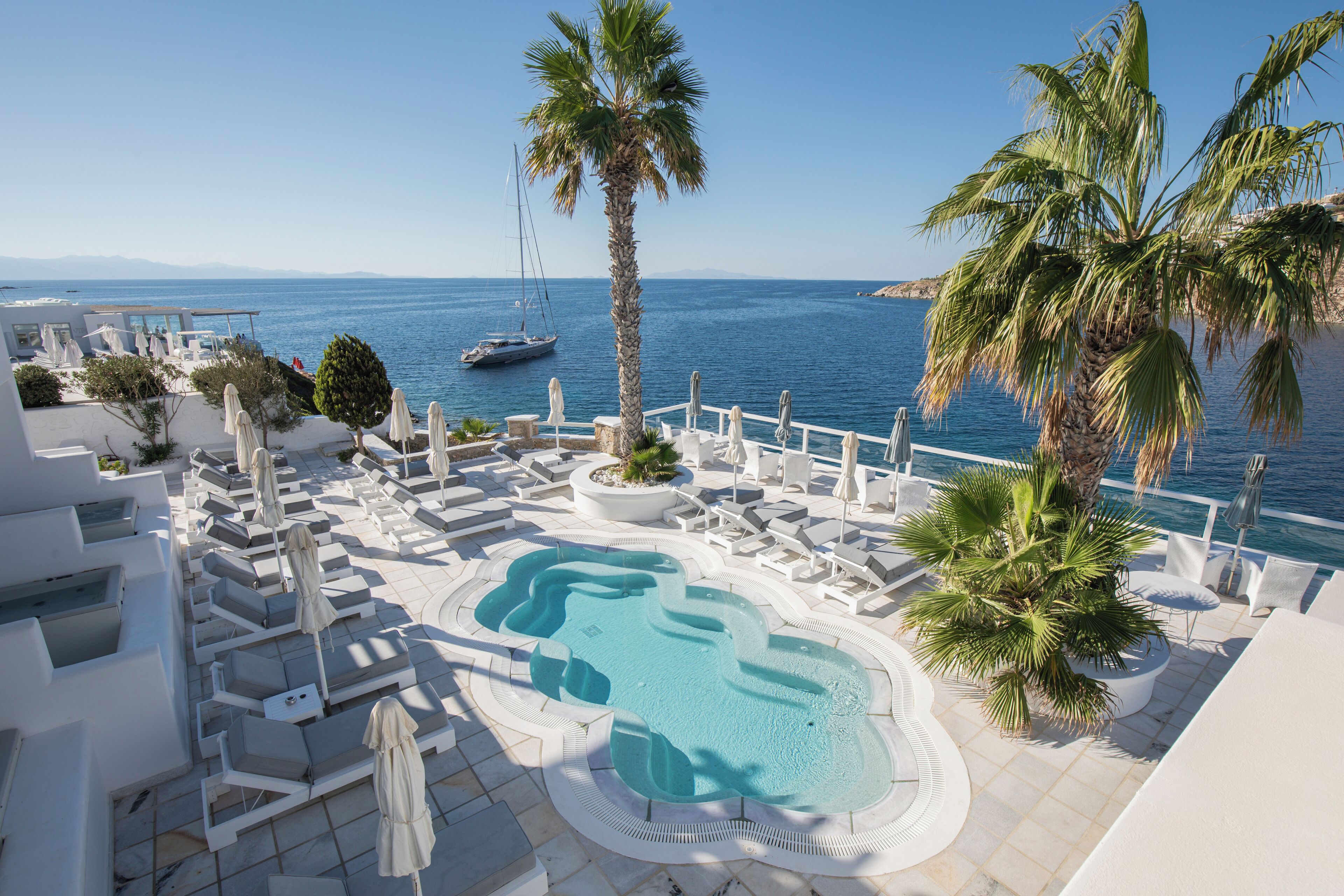 Un espace piscine avec des chaises longues et des parasols, un jacuzzi et une vue sur la mer, avec un voilier à l'horizon.
