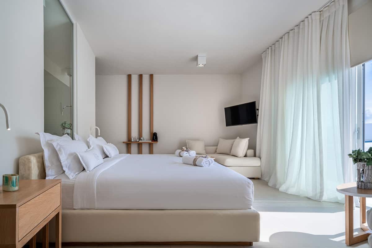 Ein modernes Hotelzimmer mit einem großen Bett, einer Sitzecke mit Sofa und Sesseln, einem flachbildfernseher an der Wand und einem großen Fenster mit Vorhängen.