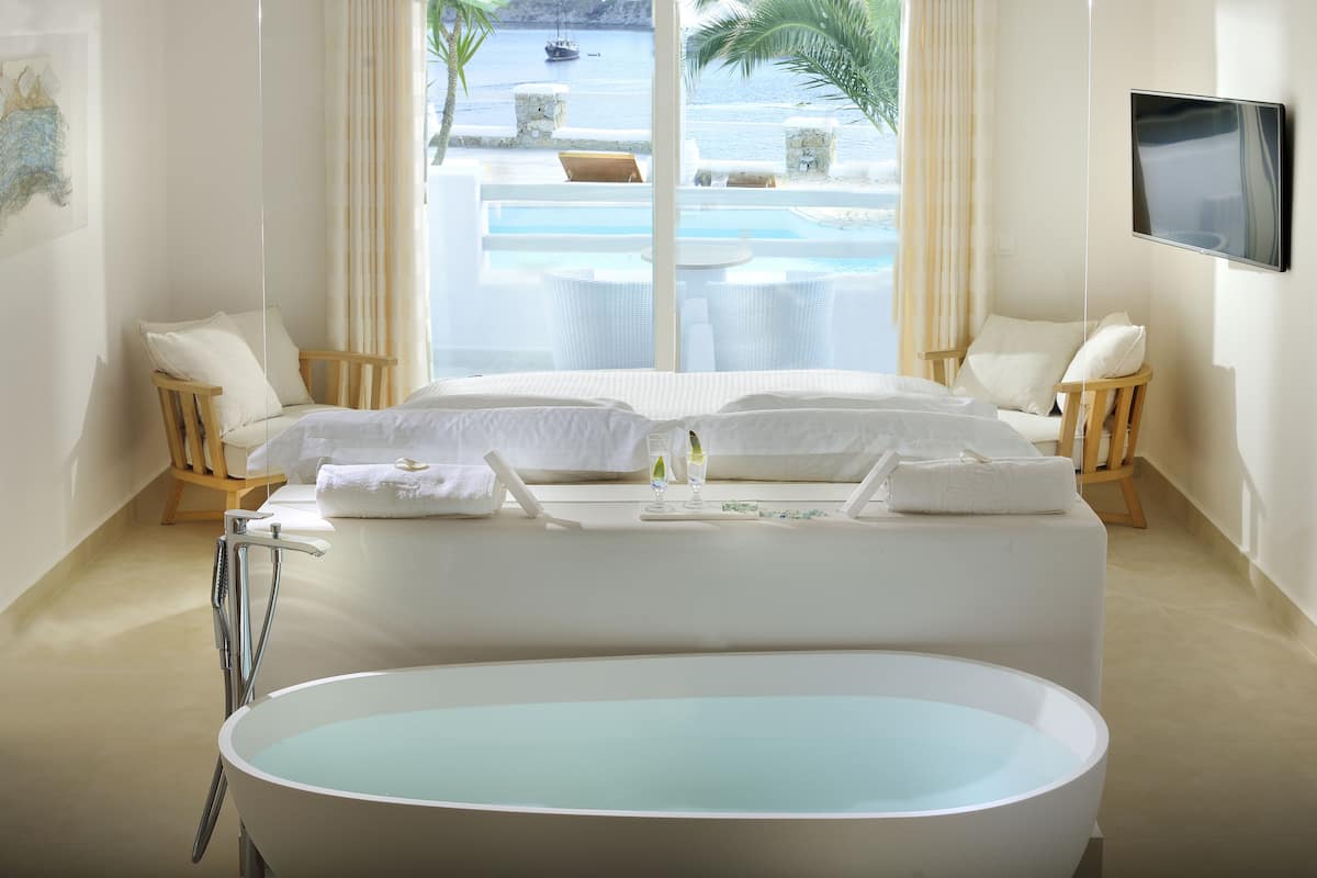 Ein modernes Hotelzimmer mit einer großen Badewanne, einem Bett mit weißer Bettwäsche, einem an der Wand montierten Fernseher und Blick auf Pool und Meer durch große Fenster.