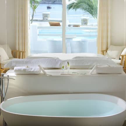 A modern hotel room with a large bathtub, a bed with white linens, a TV mounted on the wall, and a view of a pool and ocean through large windows.