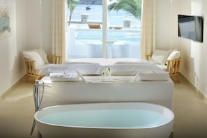 Ein modernes Hotelzimmer mit einer großen Badewanne, einem Bett mit weißer Bettwäsche, einem an der Wand montierten Fernseher und Blick auf Pool und Meer durch große Fenster.