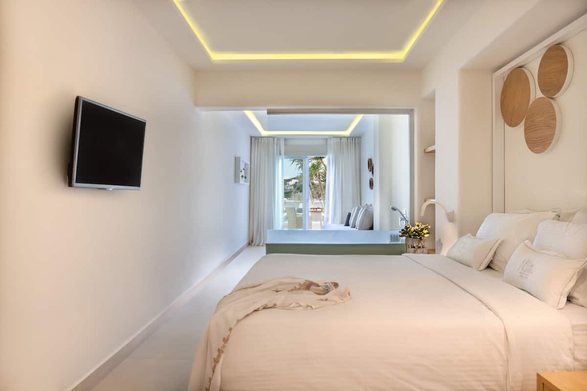 Ein modernes Schlafzimmer mit einem großen Bett, einem flachbildfernseher an der Wand und Blick ins Freie durch eine Glastür.