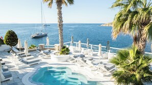 A pool area with lounge chairs and umbrellas, a palm tree, and a view of the sea with a sailboat. - Nissaki Boutique Hotel (Mykonos)