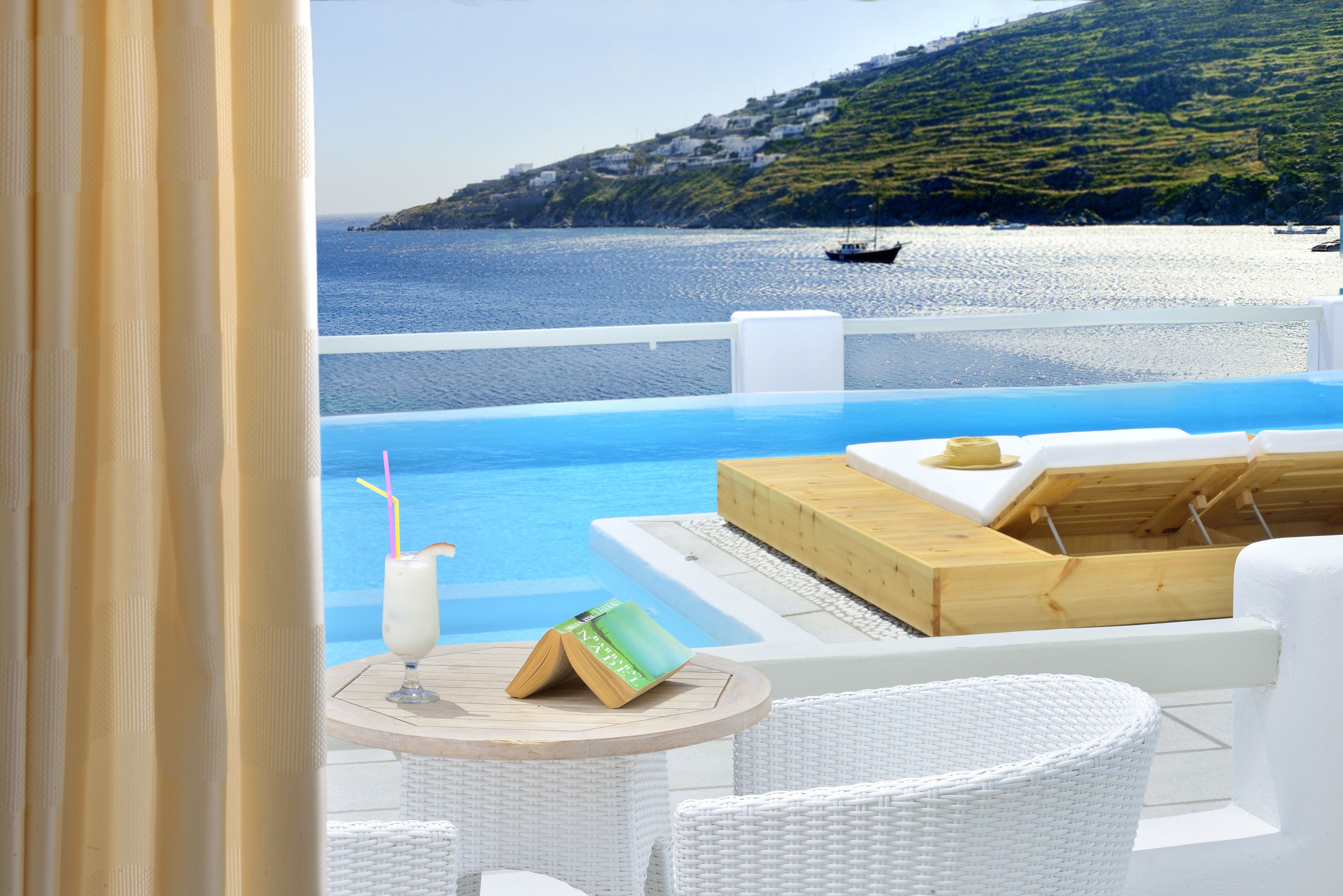 A balcony with a view of the sea, a wooden table with a book and a drink, and white wicker furniture.