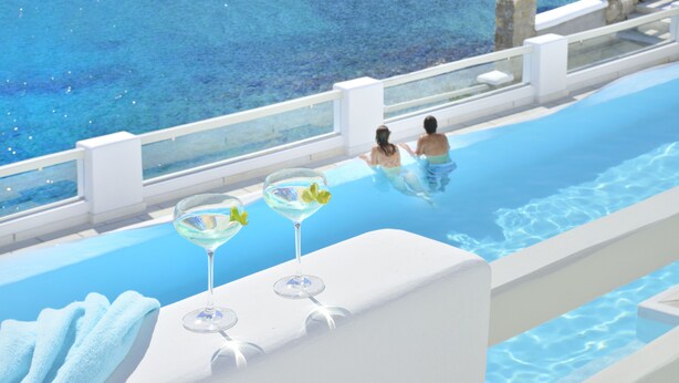 Una piscina con vista despejada al agua, zona de borde blanco y dos vasos vacíos sobre una superficie blanca.
